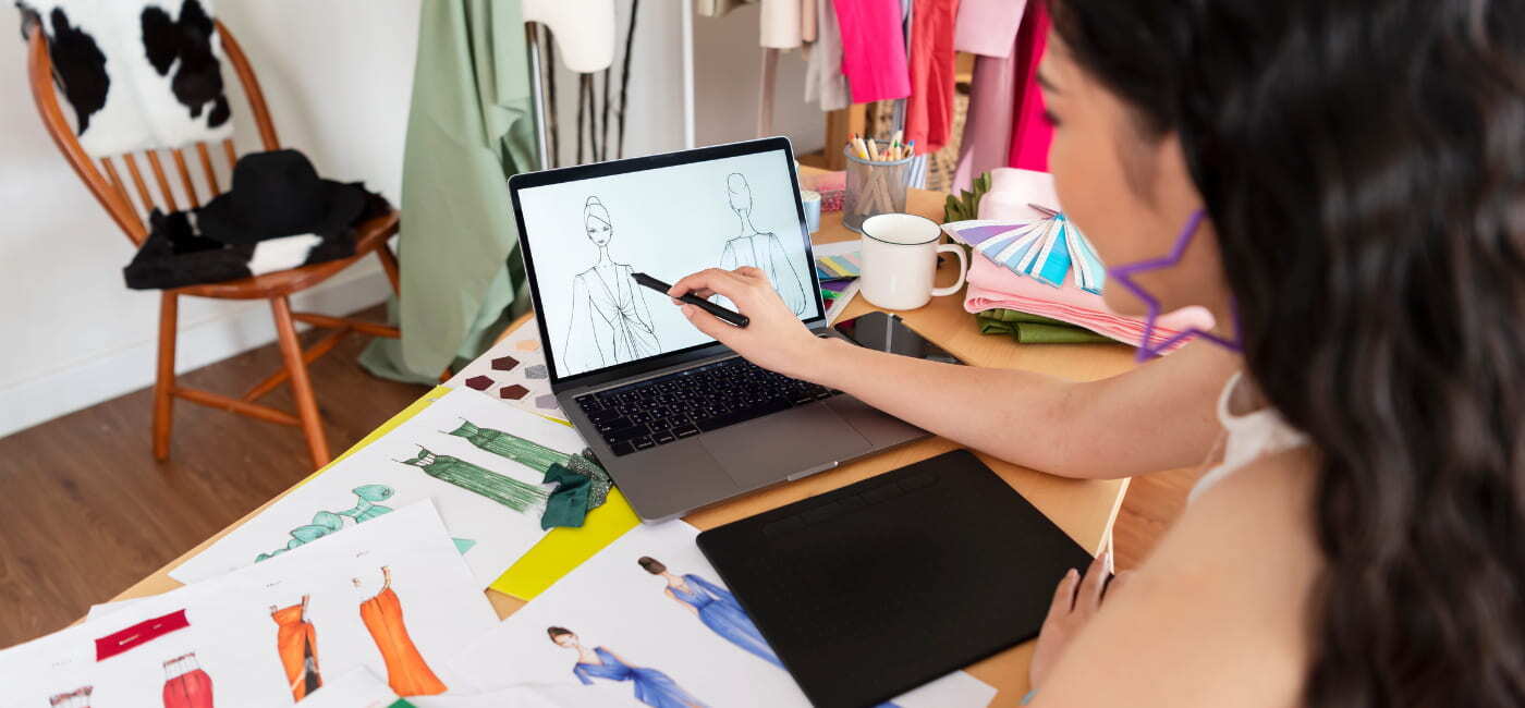 Estudiante de diseño de modas creando bocetos digitales en una laptop rodeada de telas y muestras de color.