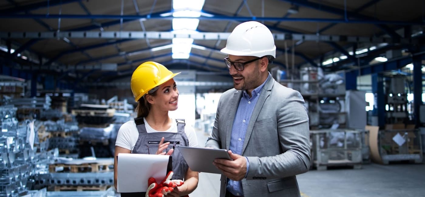 Dos profesionales, un gerente o ingeniero con traje, casco blanco y tableta, y una técnica o supervisora con overol, casco amarillo y portapapeles, conversan y sonríen dentro de una nave industrial o fábrica. La imagen representa la dirección o gestión de operaciones en la ingeniería industrial.