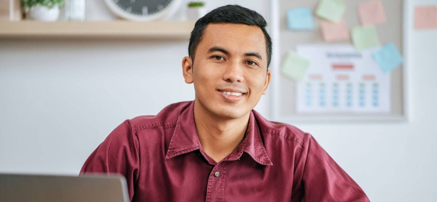 Primer plano de un joven profesional o estudiante sonriente, con camisa de botones color vino, sentado frente a un escritorio con una laptop y un tablero de planificación con post-its y un calendario al fondo.