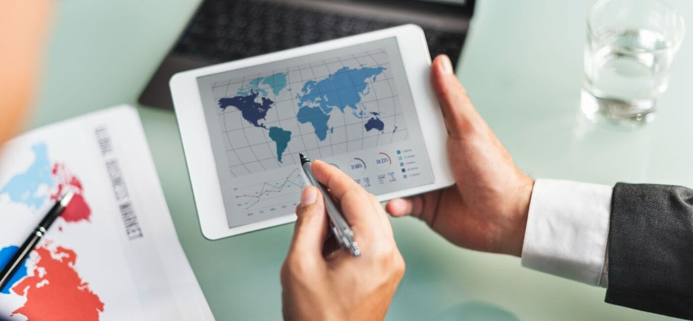 Primer plano de una persona de negocios en traje revisando un gráfico de análisis de datos y un mapa mundial en una tableta, con documentos adicionales sobre el 'Global Business Market' sobre la mesa, representando la naturaleza internacional de los negocios.