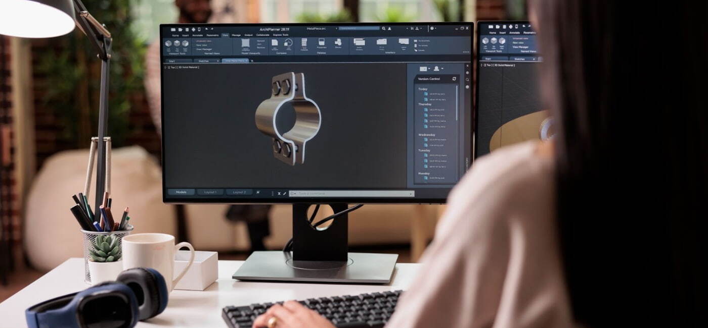 Primer plano de una diseñadora industrial trabajando en un monitor con un diseño 3D de una pieza metálica o soporte mecánico en un software CAD, con la espalda hacia la cámara.