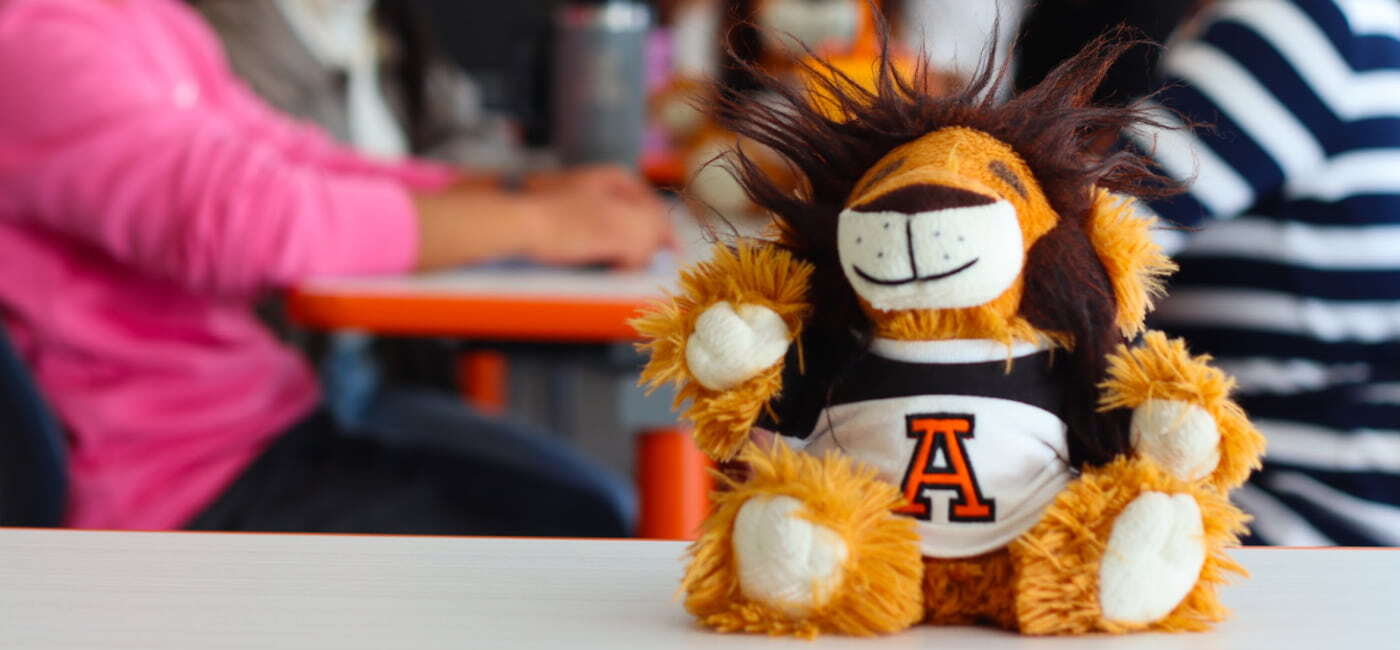 Peluche de león con camiseta de la Universidad Anáhuac sobre una mesa, con estudiantes desenfocados al fondo.